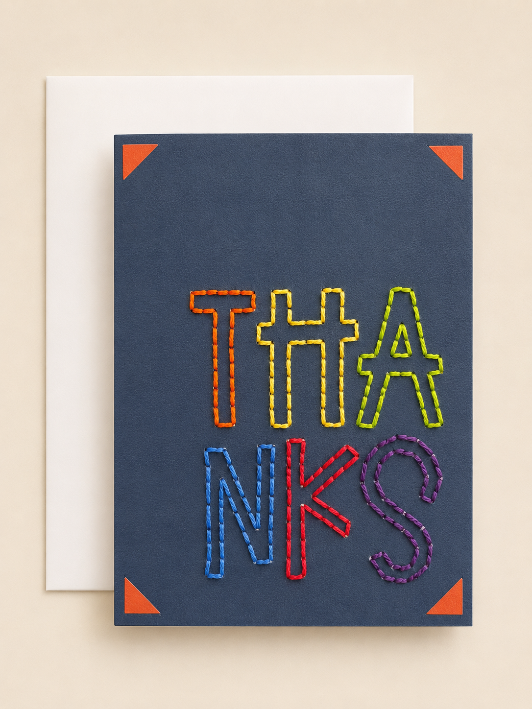 A square navy blue greeting card with the word 'THANKS' spelled out in colorful, thread-like letters, shown with a white envelope behind it on a warm beige background.