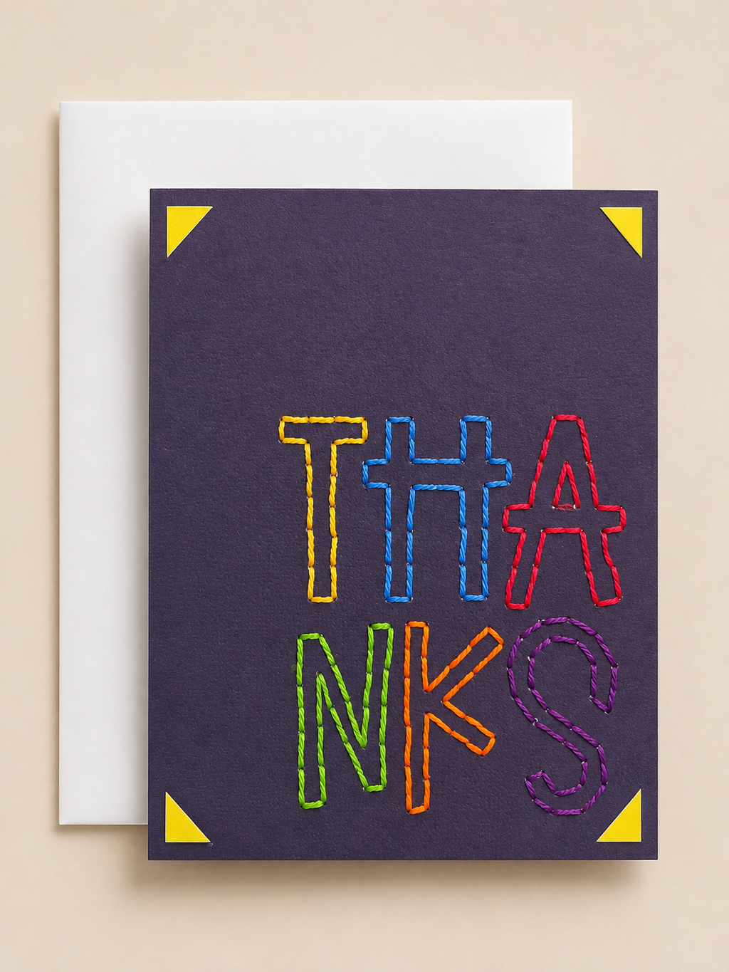 A square greeting card with a dark purple background and the word 'THANKS' in large, colorful outlined letters, shown with a white envelope behind it on a beige background.