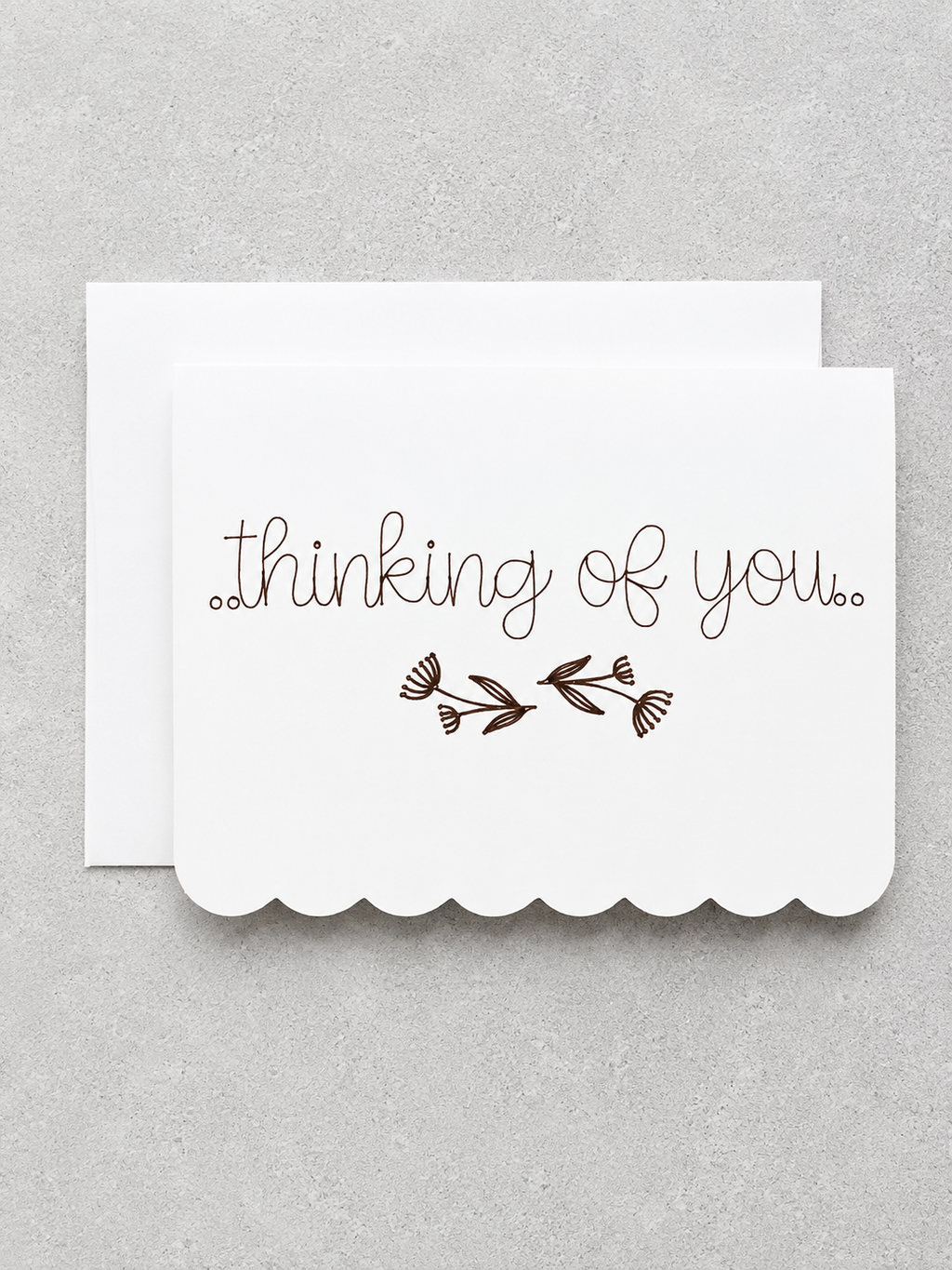 A white scalloped greeting card with the words 'thinking of you.' in brown cursive and a small brown floral motif, shown with a matching white envelope on a light gray textured background.