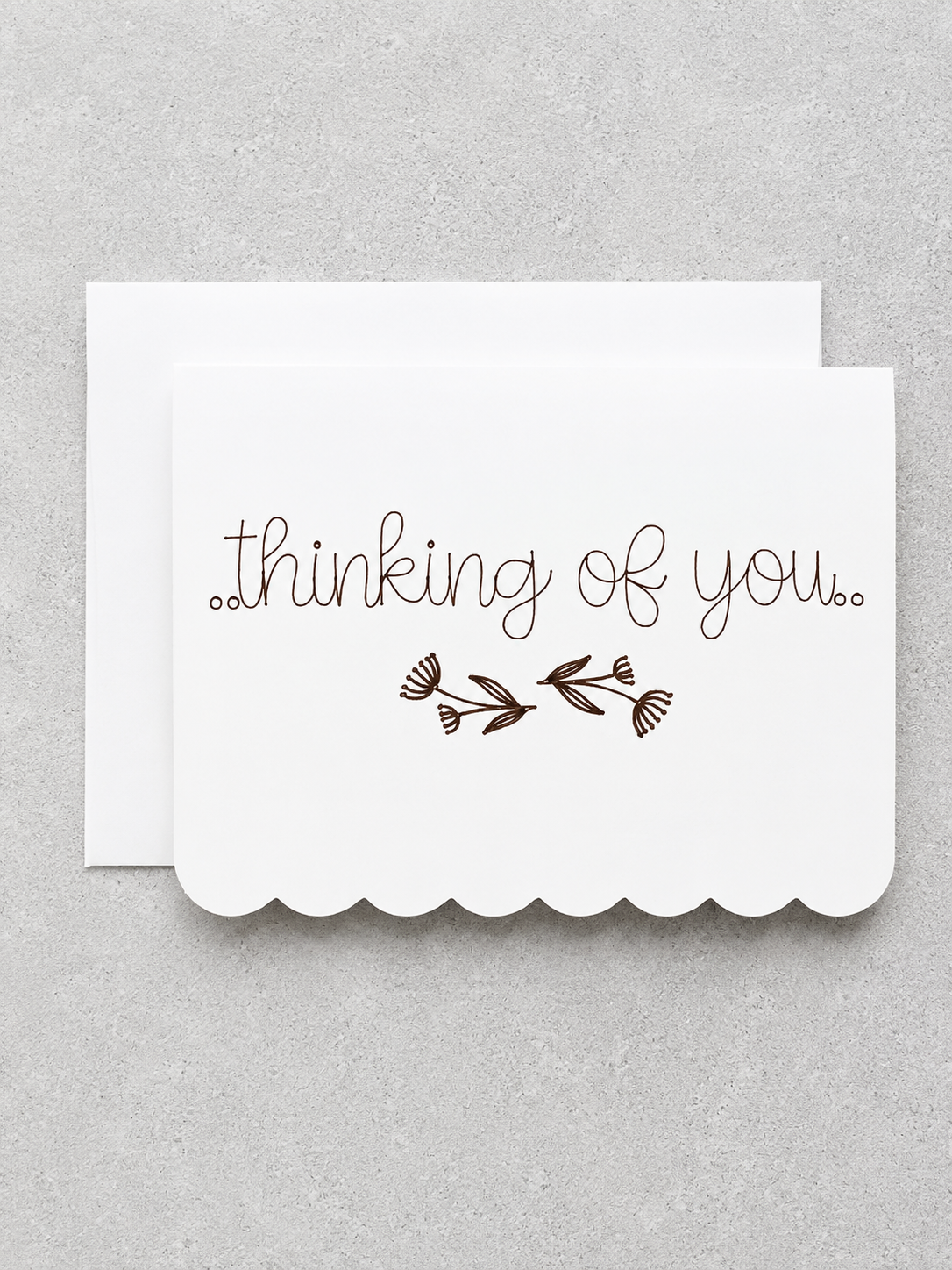 A white scalloped greeting card with the words 'thinking of you.' in brown cursive and a small brown floral motif, shown with a matching white envelope on a light gray textured background.
