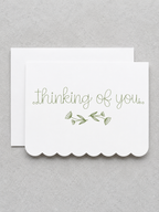 A white scalloped-edge greeting card with the words 'thinking of you.' in olive green script and a small green floral sprig, shown with a matching white envelope on a light gray textured background.
