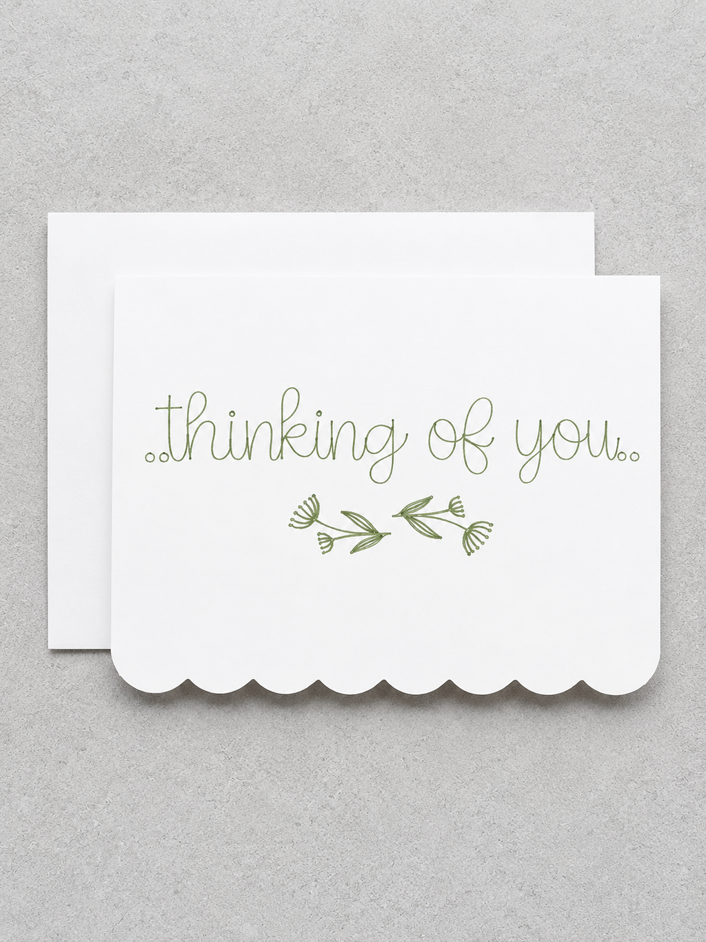 A white scalloped-edge greeting card with the words 'thinking of you.' in olive green script and a small green floral sprig, shown with a matching white envelope on a light gray textured background.