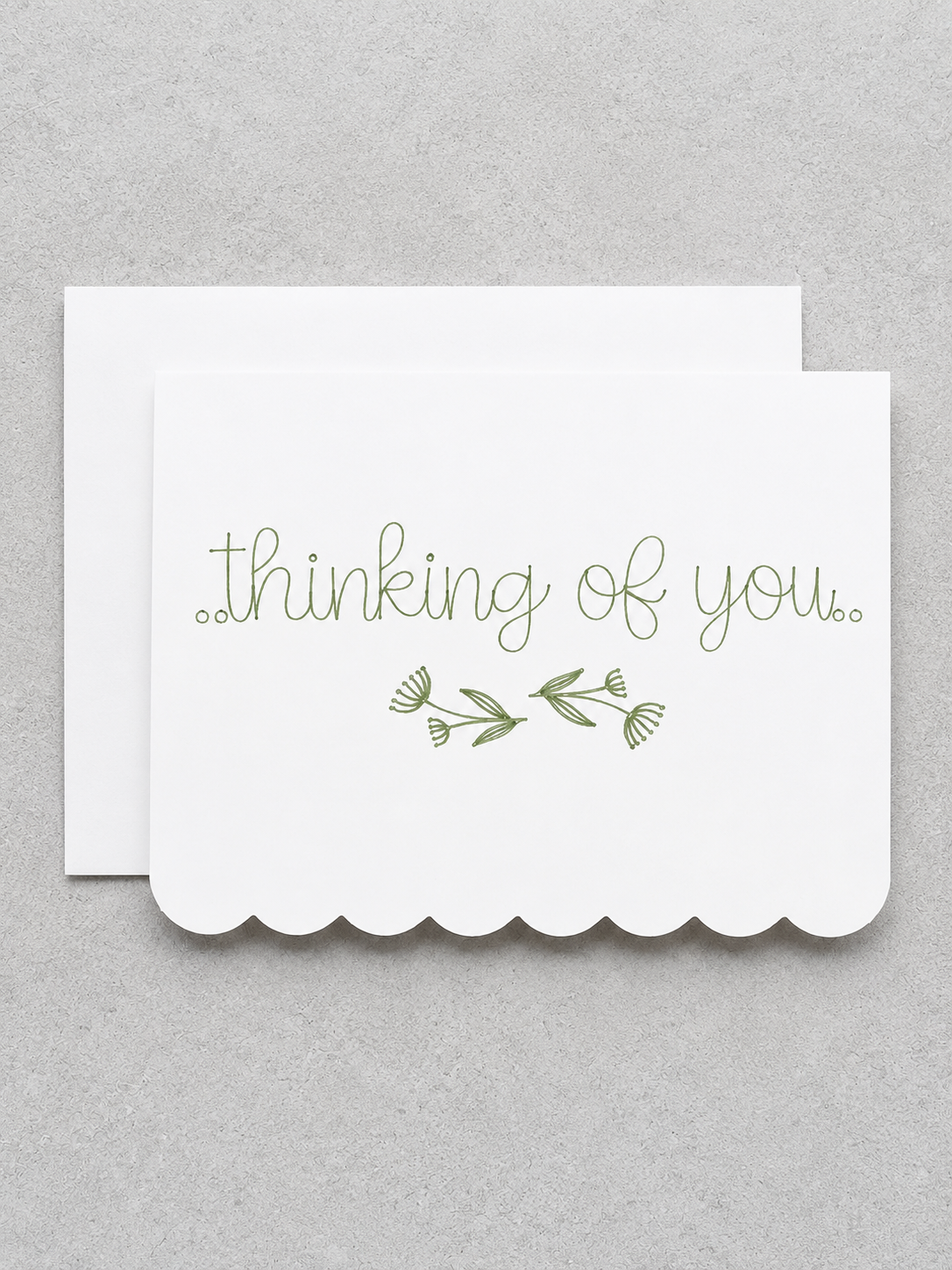 A white scalloped-edge greeting card with the words 'thinking of you.' in olive green script and a small green floral sprig, shown with a matching white envelope on a light gray textured background.