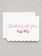 A white scalloped-edge greeting card with the words 'thinking of you.' in red cursive script and a small red floral branch motif, shown with a matching white envelope on a light gray textured background.