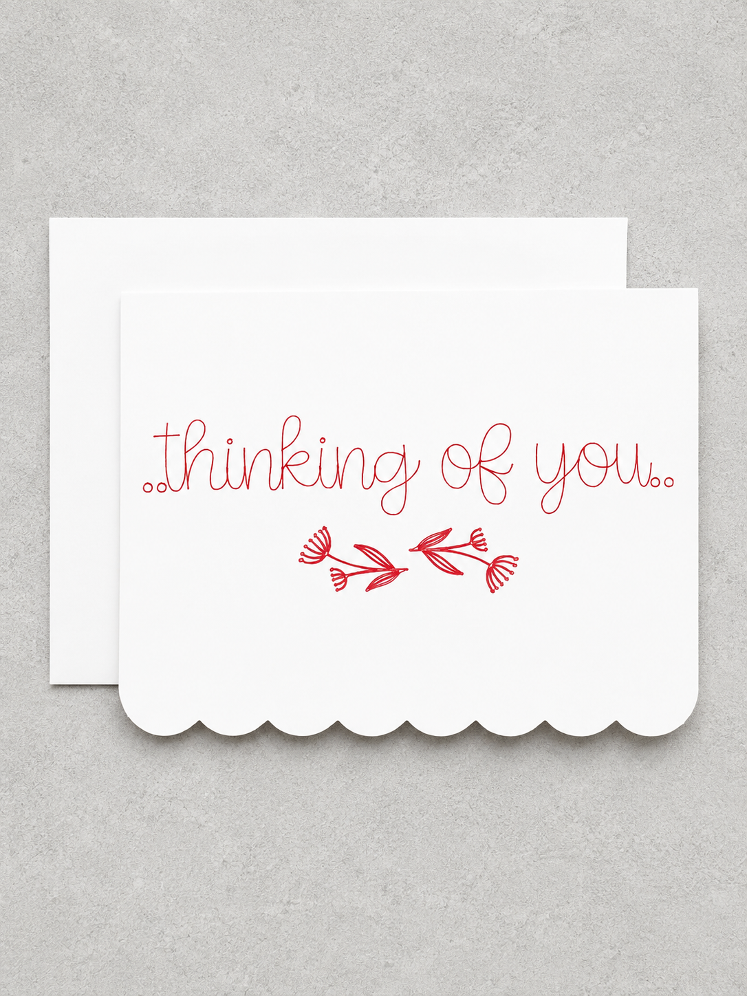 A white scalloped-edge greeting card with the words 'thinking of you.' in red cursive script and a small red floral branch motif, shown with a matching white envelope on a light gray textured background.