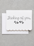 A white scalloped-edge greeting card with the words 'thinking of you.' and a small black floral illustration, shown with a matching white envelope on a textured gray background.