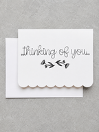 A white scalloped-edge greeting card with the words 'thinking of you.' and a small black floral illustration, shown with a matching white envelope on a textured gray background.