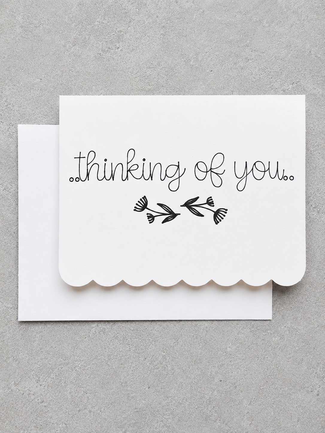 A white scalloped-edge greeting card with the words 'thinking of you.' and a small black floral illustration, shown with a matching white envelope on a textured gray background.