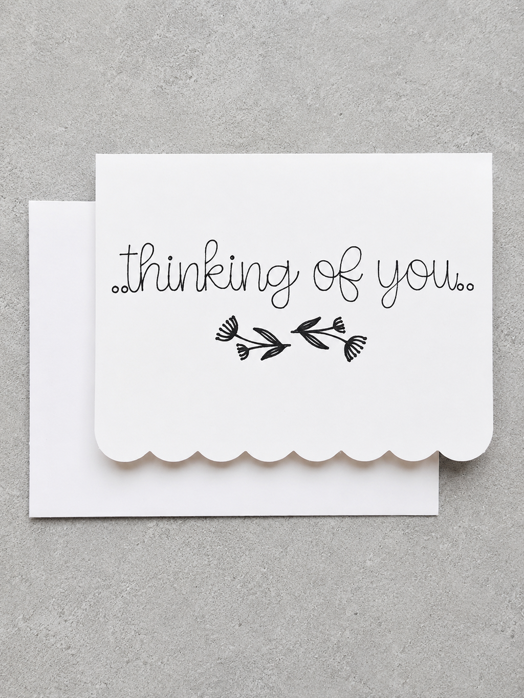 A white scalloped-edge greeting card with the words 'thinking of you.' and a small black floral illustration, shown with a matching white envelope on a textured gray background.