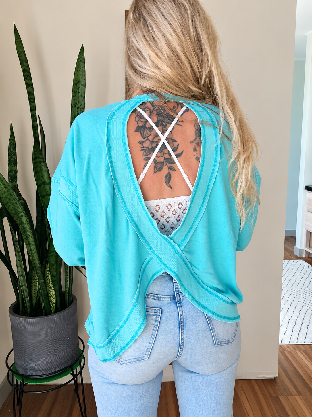 A woman viewed from behind wearing a turquoise long-sleeve wrap-style top with a large open back and white crisscross straps, paired with light blue jeans. She stands indoors next to a potted snake plant.
