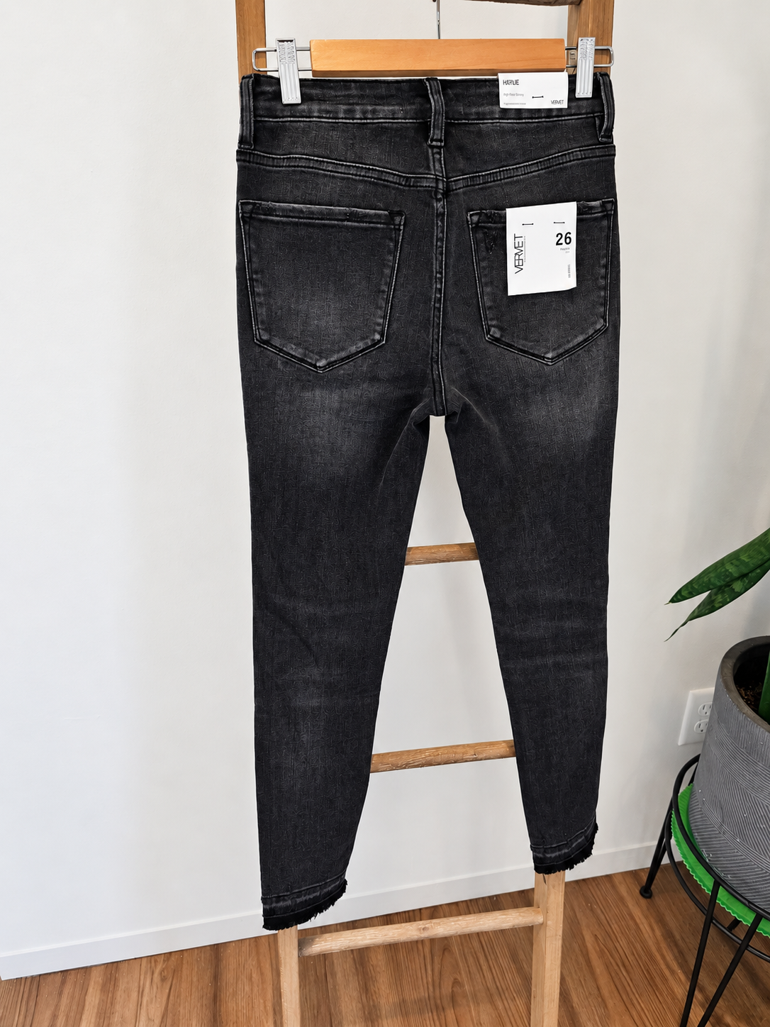 Back view of a pair of dark gray washed skinny jeans hanging on a wooden hanger, displayed on a wooden step stool in an indoor setting.