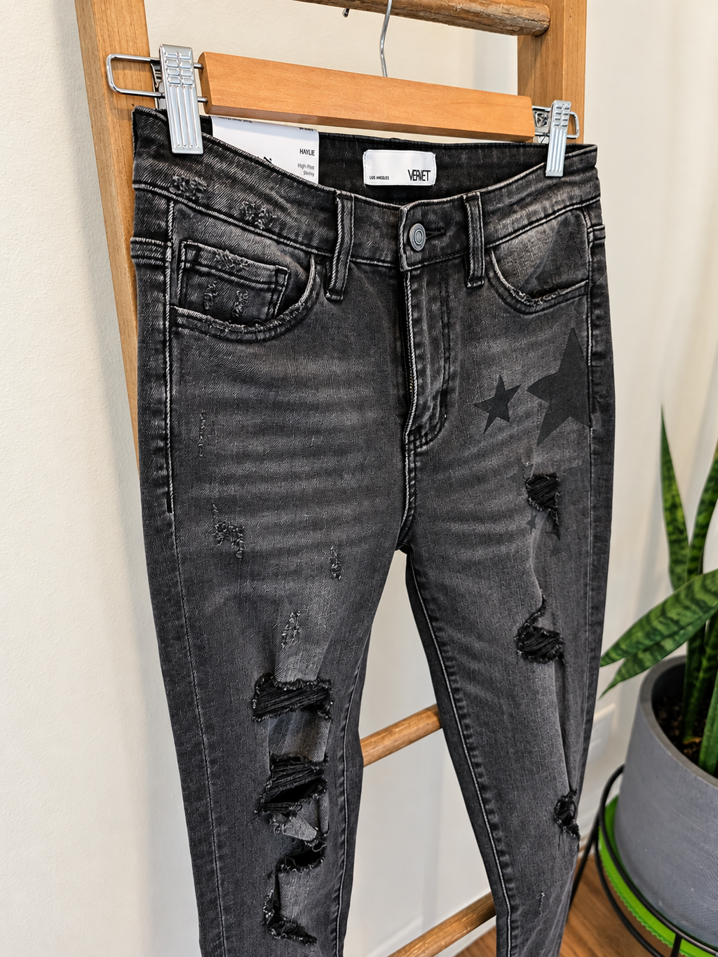 Black washed distressed skinny jeans with ripped knees and star graphics on the thighs, hanging on a wooden hanger on a wooden ladder indoors.