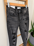 Black washed distressed skinny jeans with ripped knees and star graphics on the thighs, hanging on a wooden hanger on a wooden ladder indoors.