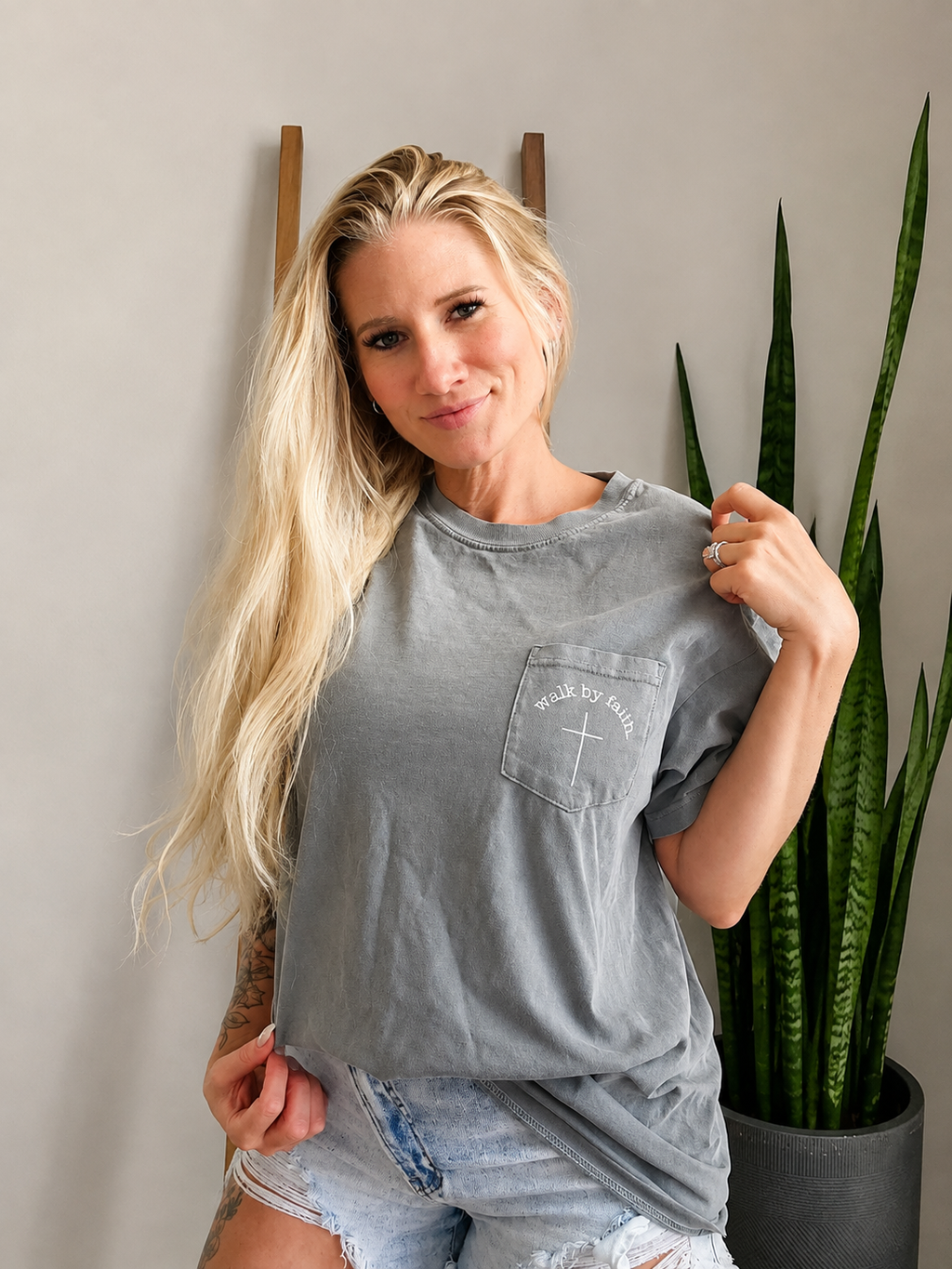 A woman with long blonde hair wearing a gray washed graphic t-shirt and denim shorts, posing in front of a gray wall with a snake plant in a pot.