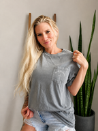 A woman with long blonde hair wearing a gray washed graphic t-shirt and denim shorts, posing in front of a gray wall with a snake plant in a pot.