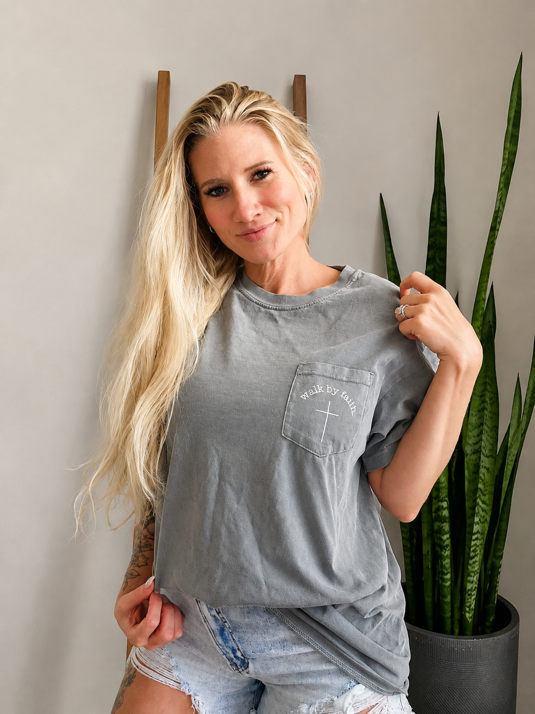A woman with long blonde hair wearing a gray washed graphic t-shirt and denim shorts, posing in front of a gray wall with a snake plant in a pot.