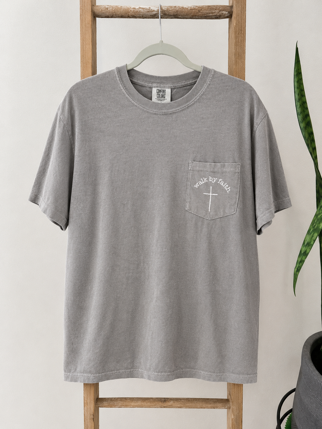 A gray short-sleeve t-shirt with a white graphic on the left chest pocket, hanging on a wooden hanger on a wooden ladder with a green plant in the background.