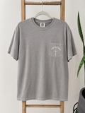 A gray short-sleeve t-shirt with a white graphic on the left chest pocket, hanging on a wooden hanger on a wooden ladder with a green plant in the background.