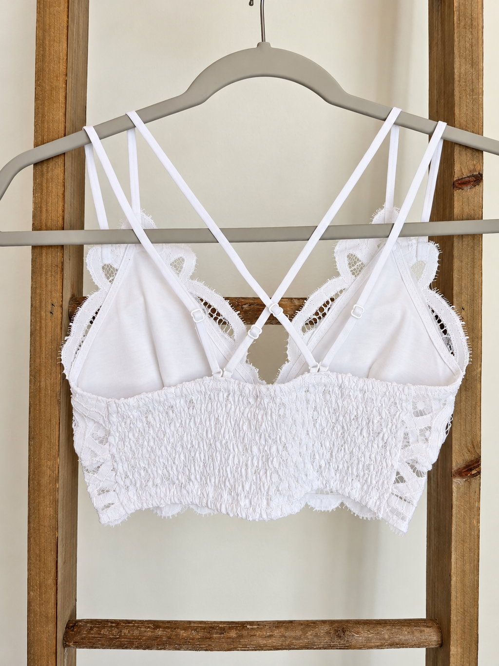 White lace and smocked crop top with thin double straps, hanging on a gray hanger against a wooden ladder background.