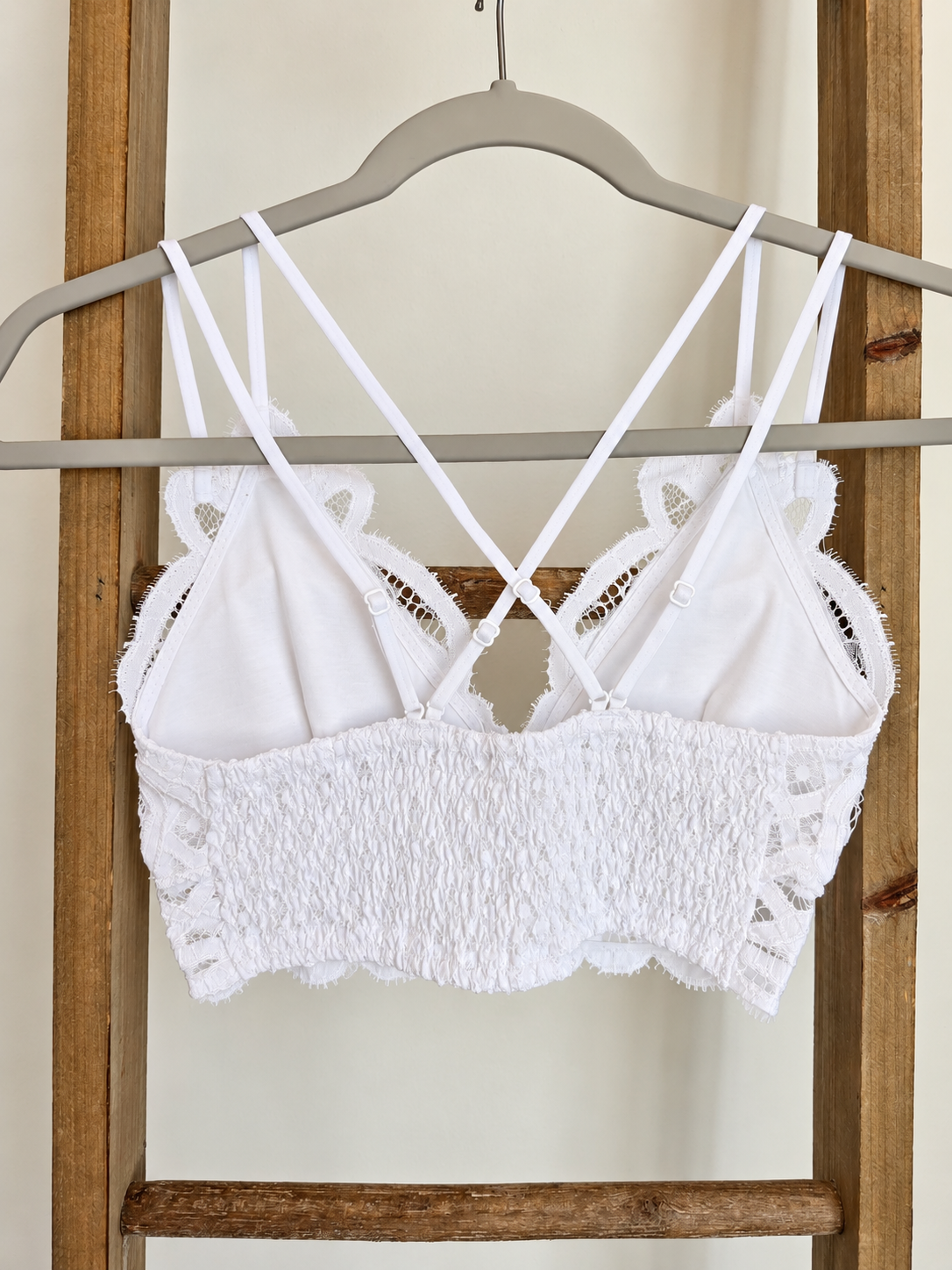 White lace and smocked crop top with thin double straps, hanging on a gray hanger against a wooden ladder background.