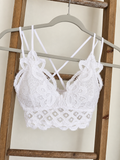White lace bralette with floral applique and thin crisscross back straps, hanging on a gray hanger against a wooden ladder backdrop.