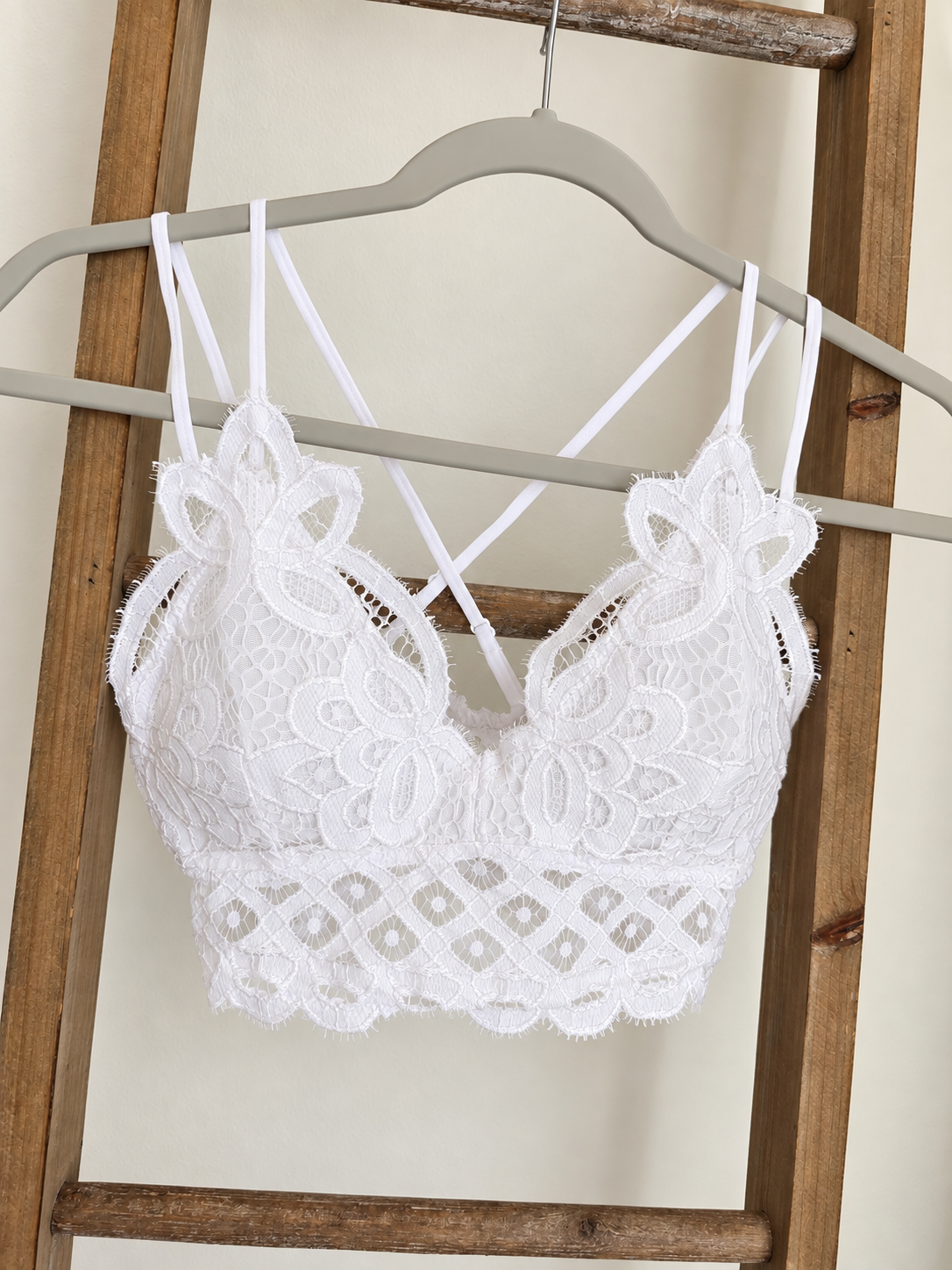 White lace bralette with floral applique and thin crisscross back straps, hanging on a gray hanger against a wooden ladder backdrop.