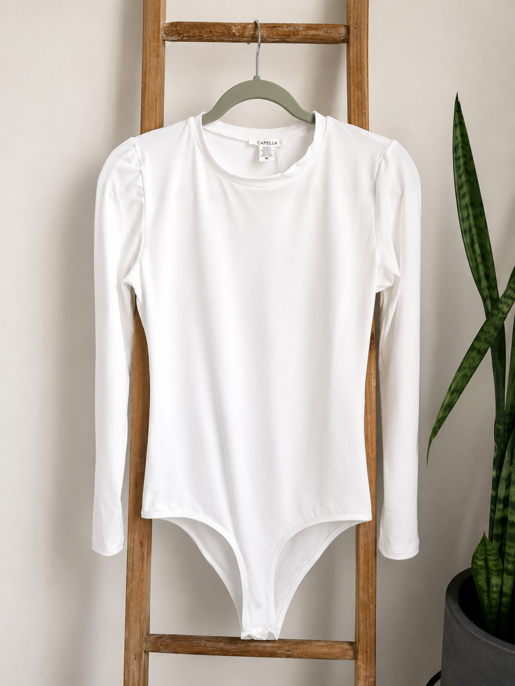 A white long-sleeve bodysuit with puffed shoulders hangs on a green hanger from a wooden ladder, with a potted snake plant visible on the right against a beige wall.