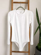 A white long-sleeve bodysuit with puffed shoulders hangs on a green hanger from a wooden ladder, with a potted snake plant visible on the right against a beige wall.