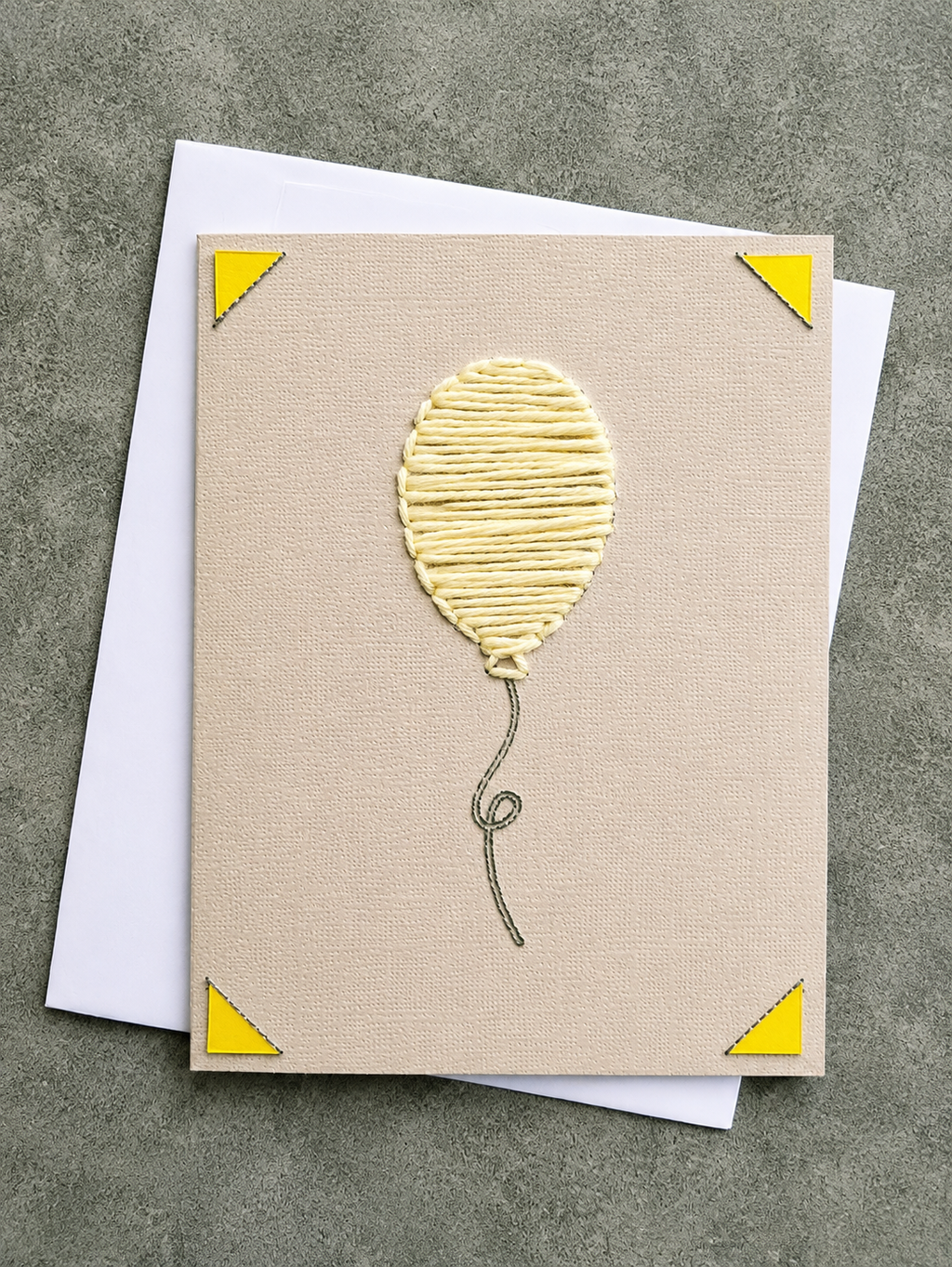 A square beige greeting card with a textured balloon design in the center, yellow corner triangles, and a gray string outline, shown with a white envelope on a gray surface.
