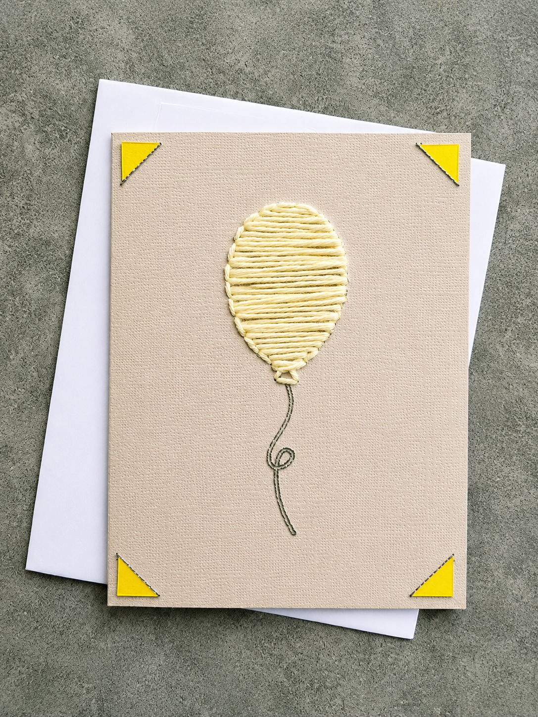 A square beige greeting card with a textured balloon design in the center, yellow corner triangles, and a gray string outline, shown with a white envelope on a gray surface.