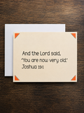 A square cream-colored greeting card with black text reading 'And the Lord said, "You are now very old." Joshua 13:1' and small orange corner accents, shown with a white envelope on a dark wood background.